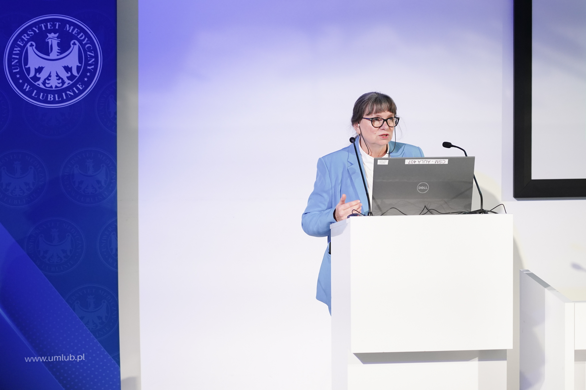 Elegancko ubrana kobieta za m&oacute;wnicą z dwoma mikrofonami, z lewej strony zdjęcia logo Uniwersytetu Medycznego w Lublinie.